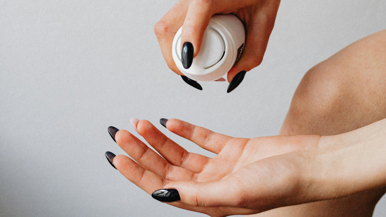 Close-up of a woman applying self tanner mousse into her hand before application, showing the texture and color of the tanning foam against pale skin and black manicured nails.