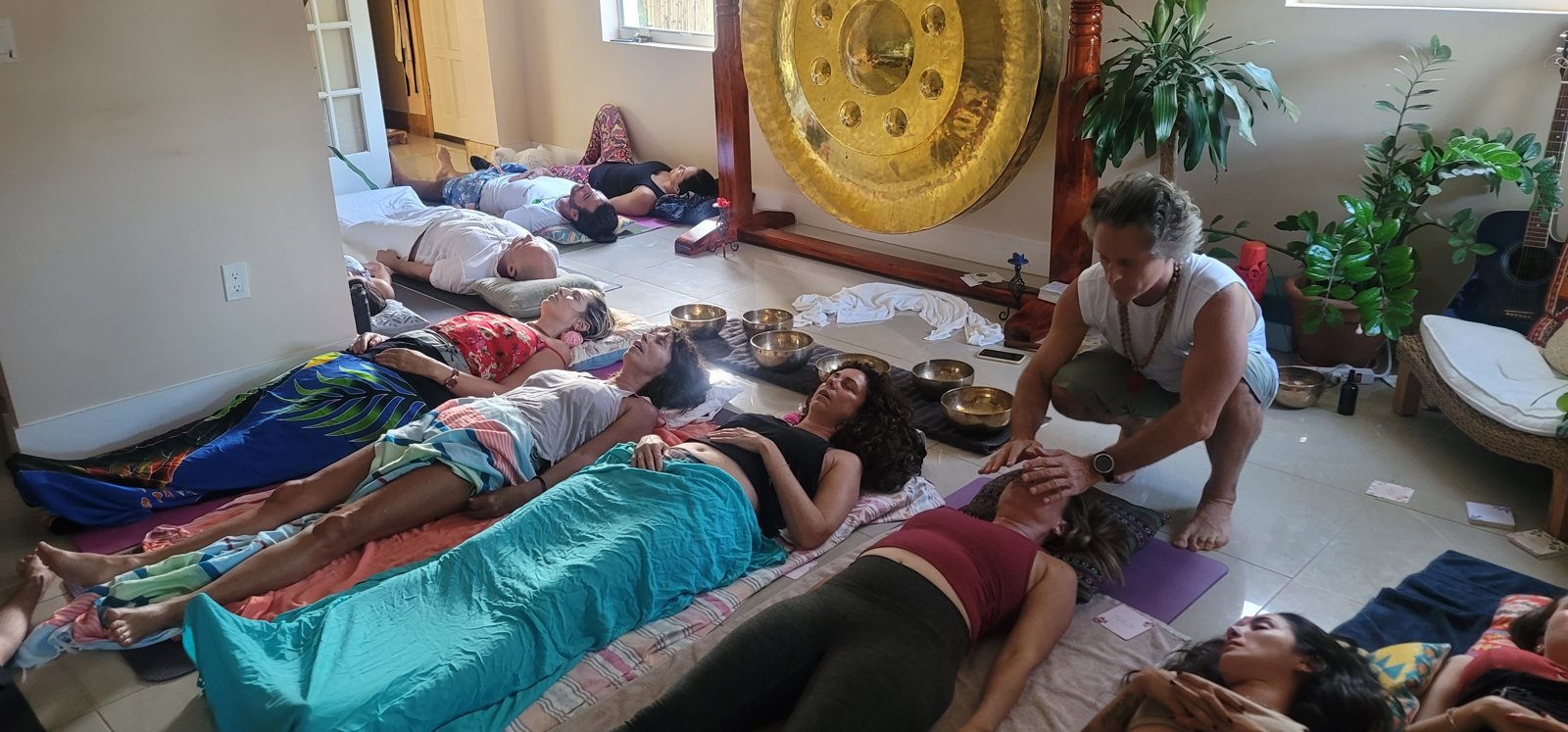Friends lying on mats in a sunlit living room at a home breathwork gathering, with a gong and singing bowls