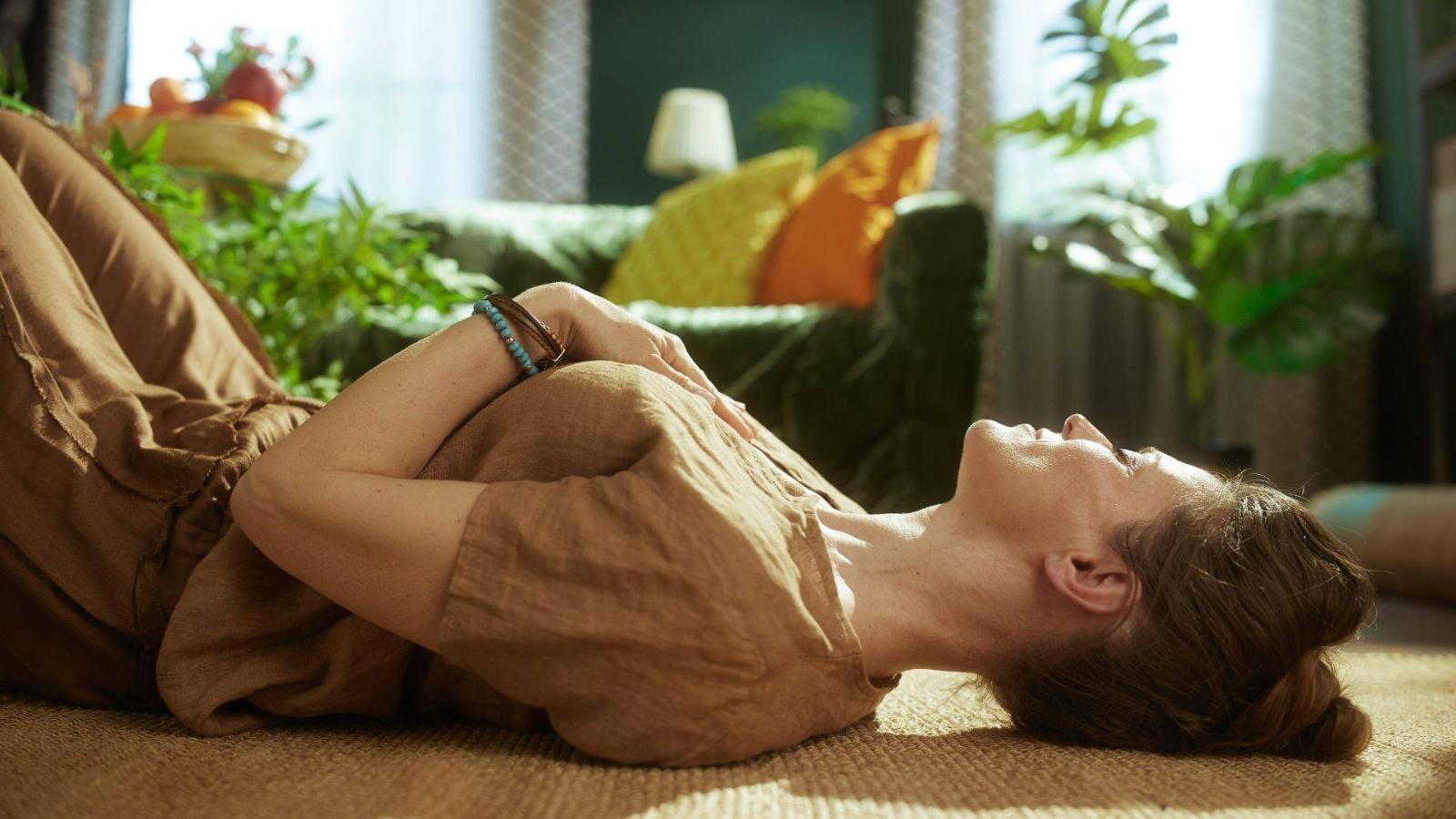 A woman lying back with her hands on her chest, breathing in a warm, plant-filled room