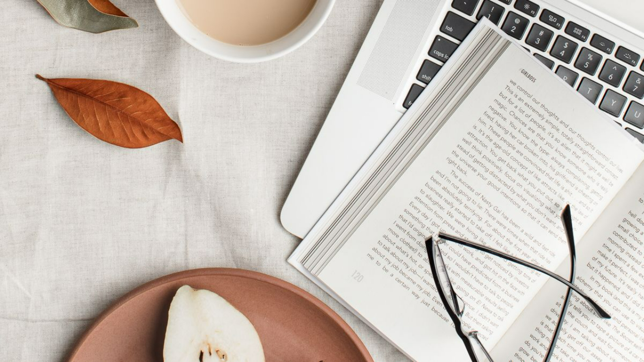 Person in a cream sweater holding a mug beside an open journal at a bright, clean desk by a window.