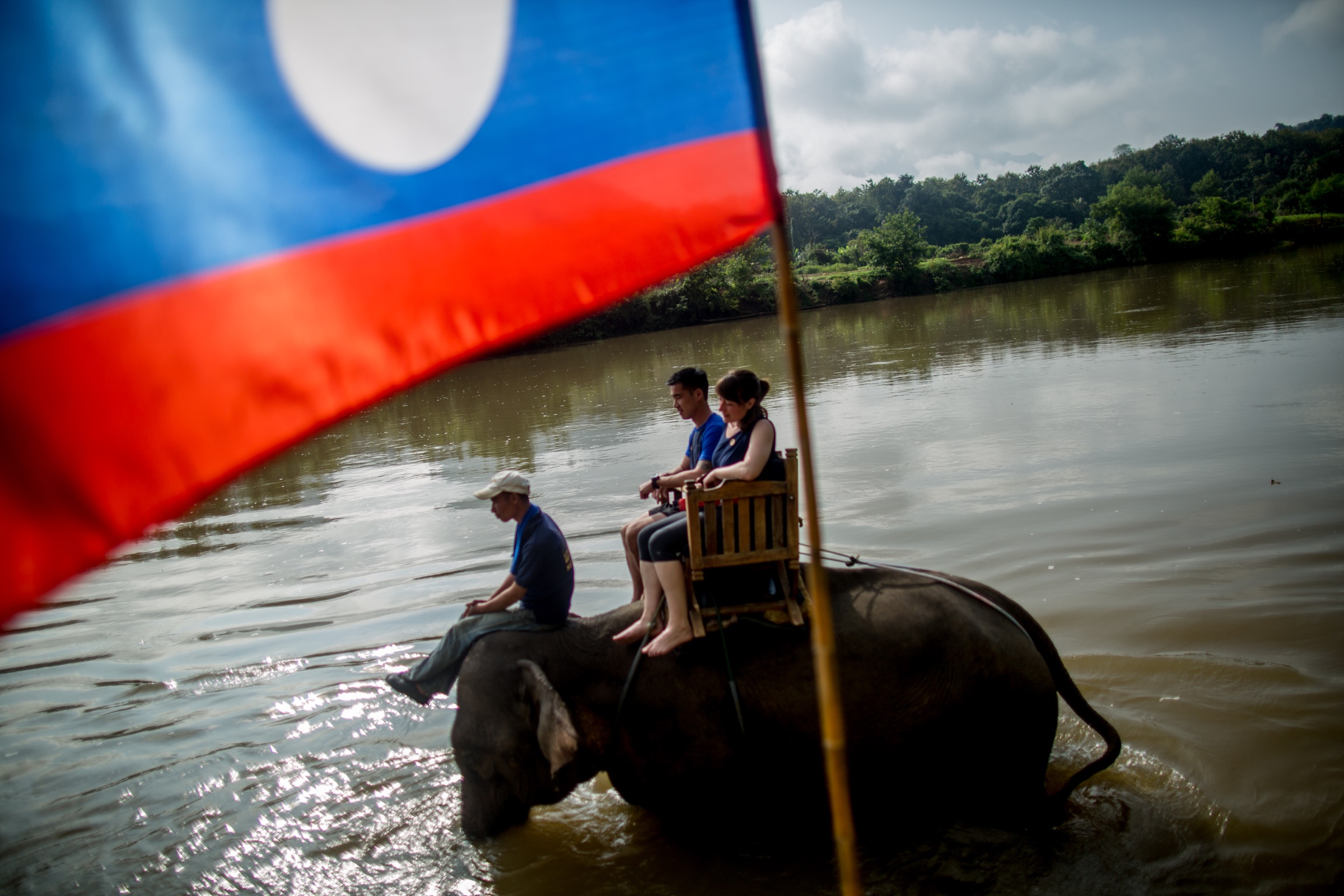 A Lao flag frames tourists and a handler riding an elephant through a river in Luang Prabang.