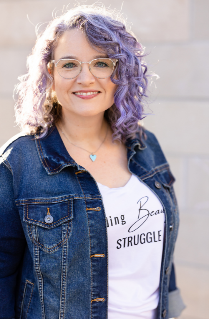 Close-up portrait of Maria smiling gently while wearing a denim jacket and white shirt, with soft purple curls and a warm natural background.