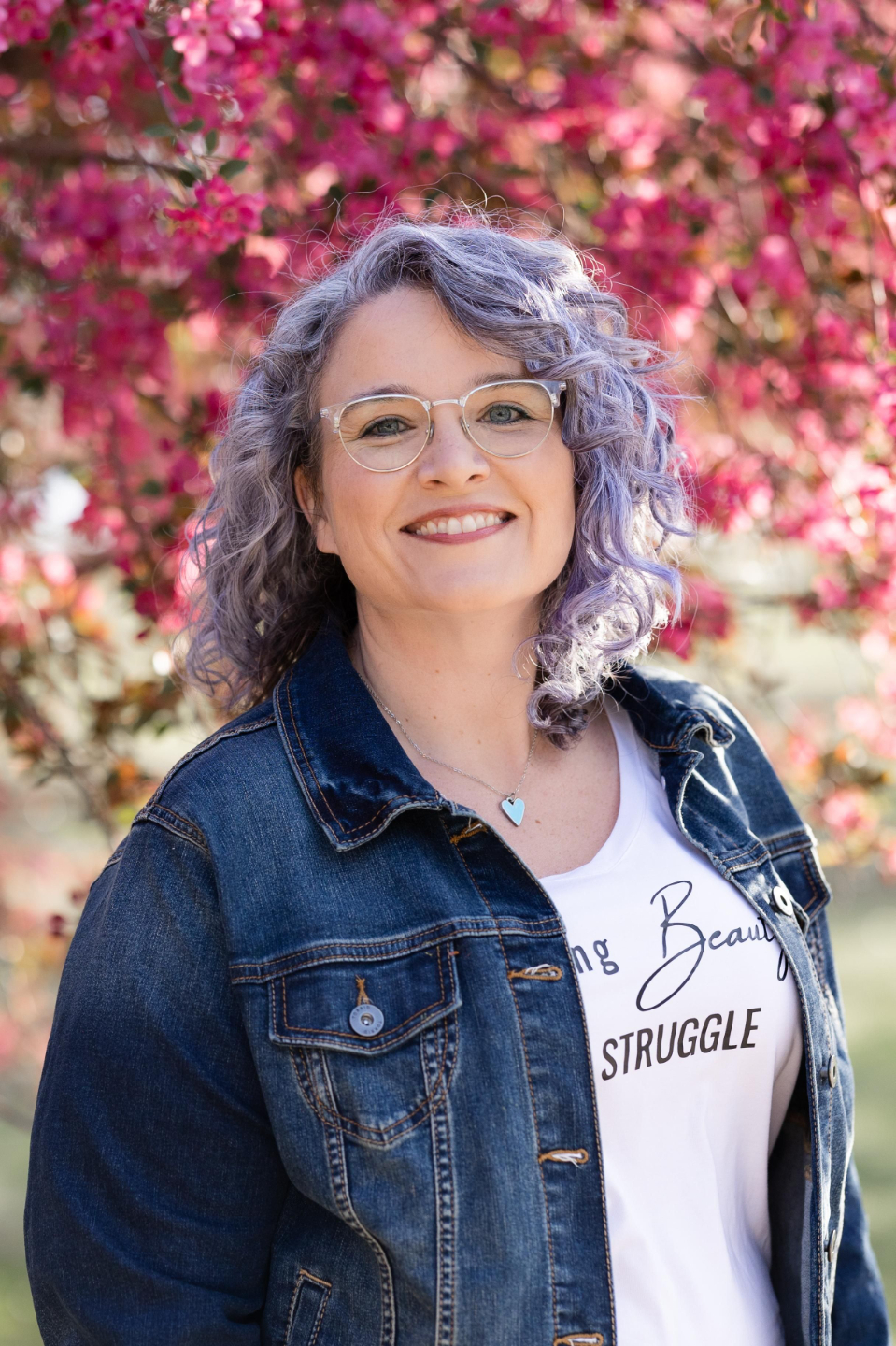 Portrait of Maria standing in front of blooming pink flowers, smiling naturally in a denim jacket and glasses with a bright and uplifting atmosphere.