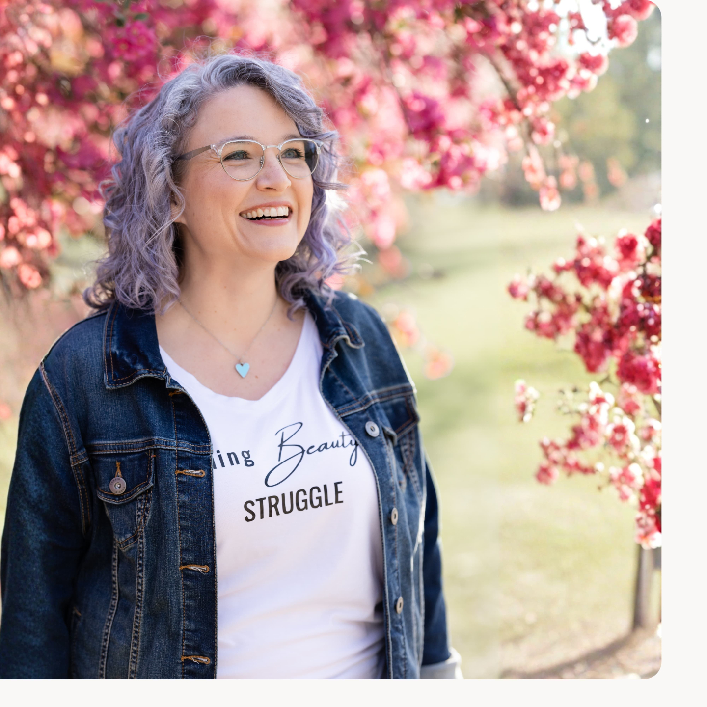 Maria smiling in a garden filled with pink blossoms, wearing a casual denim jacket and glasses, with a warm and approachable feel.