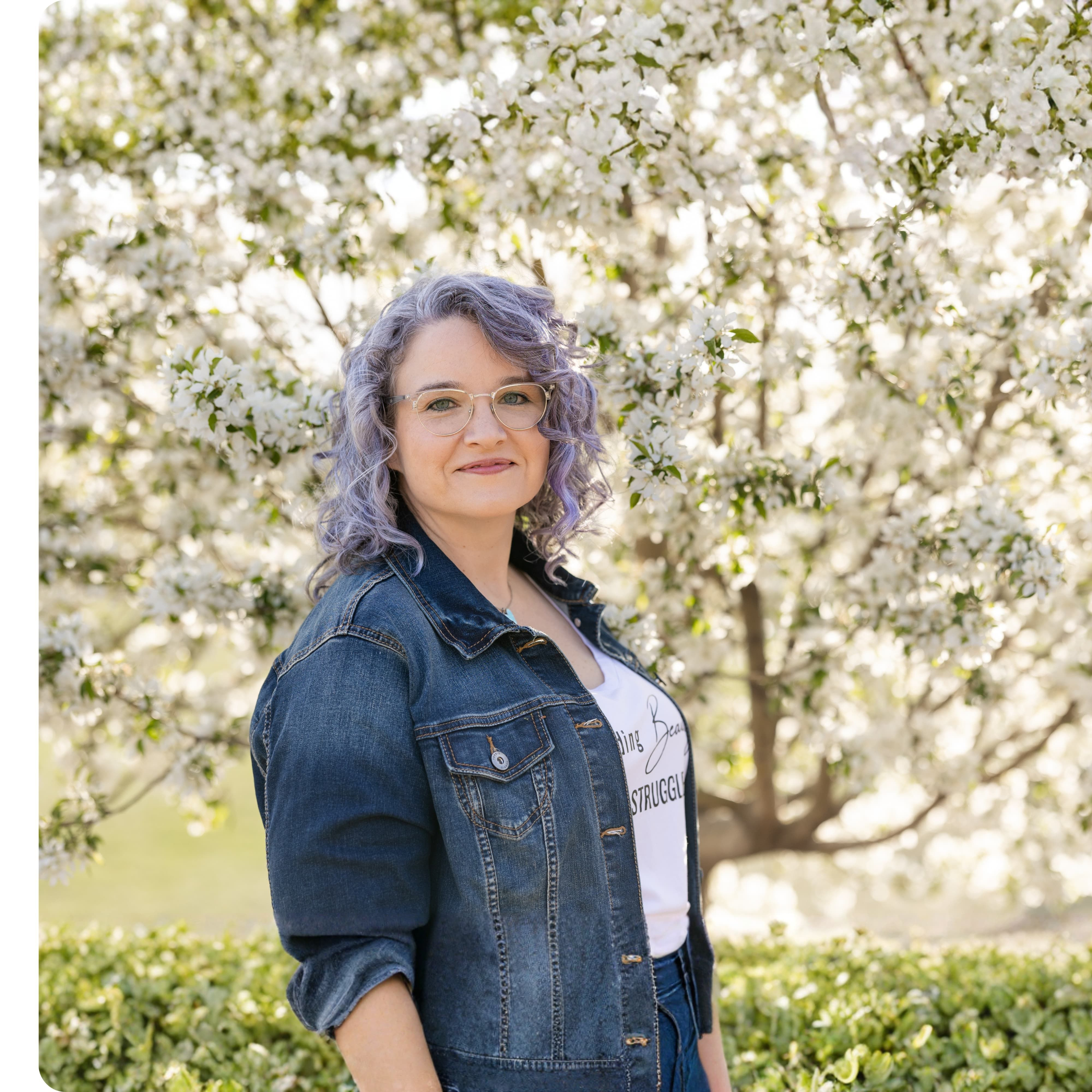 Maria posing in a garden surrounded by blooming white flowers, wearing glasses and a denim jacket, creating a soft and approachable feel.