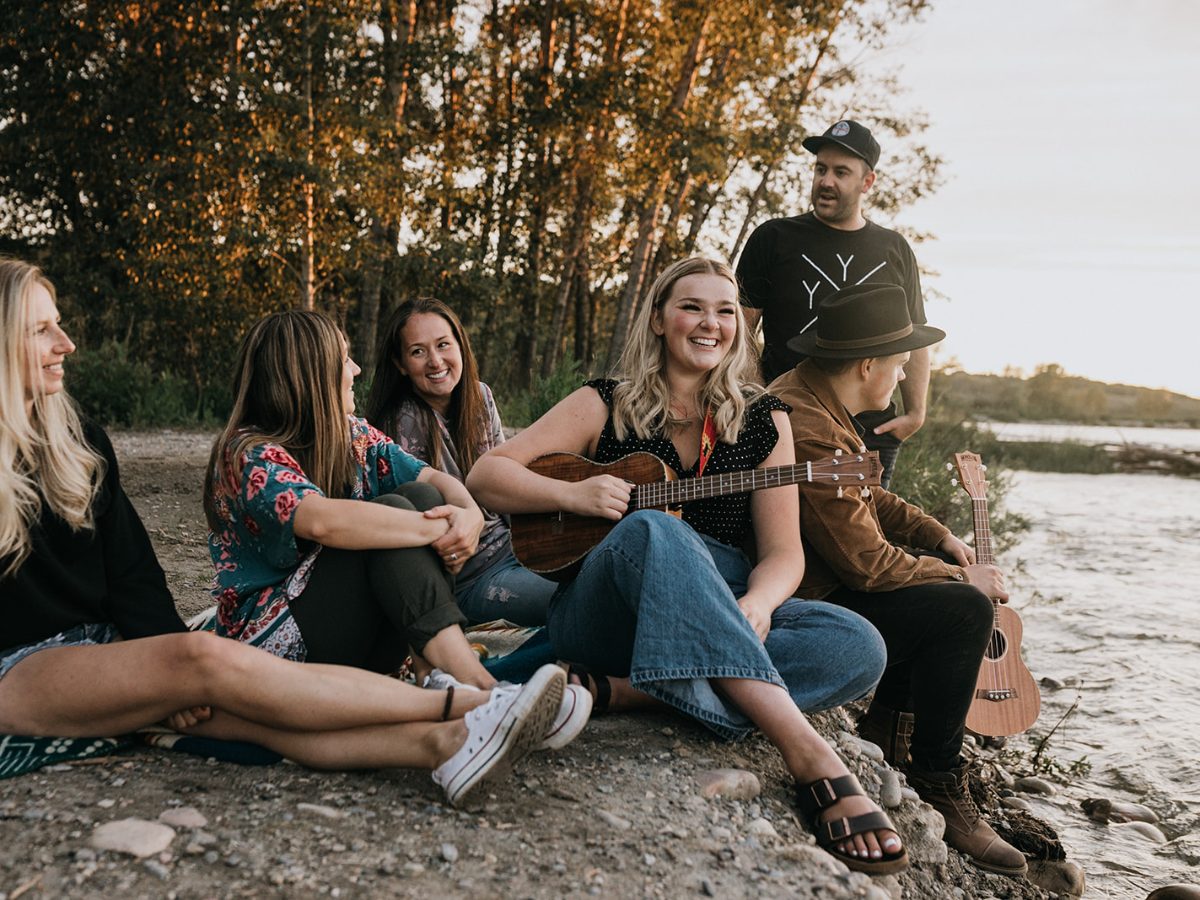 A group of friends by the river at sunset, singing along while one plays the ukulele