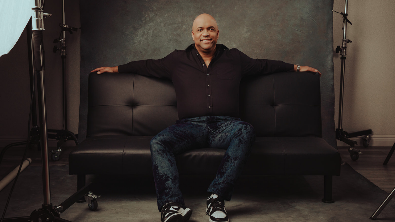 Mike Veny, mental health keynote and motivational speaker and professional drummer, smiles confidently while seated on a black couch with his arms extended in a dramatic photography studio setting, wearing a black shirt, blue patterned pants, and black-and-white sneakers.