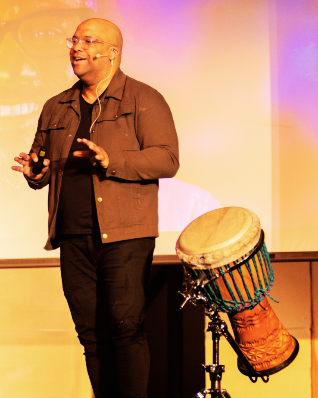 Mike Veny stands on a stage with his arms open beside a large hand drum on a stand, appearing to engage an audience during an interactive speaking or drumming presentation. He is wearing a light-colored blazer, white shirt, jeans, and sneakers, with a microphone positioned in front of the drum. The stage backdrop includes black curtains, plants, and decorative set pieces, creating a warm event atmosphere. This image highlights Mike Veny as a mental health speaker, wellness advocate, and interactive drumming facilitator.