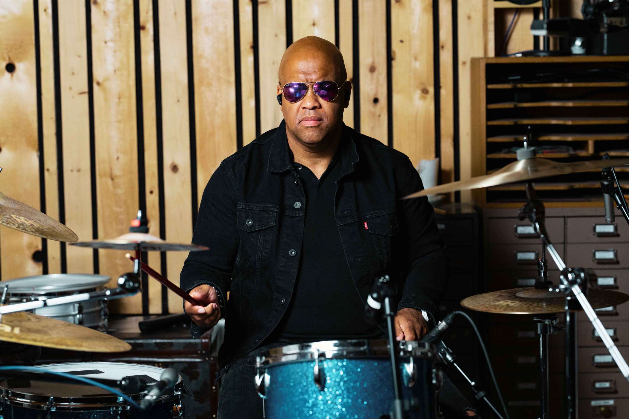 Professional drummer Mike Veny sits confidently on the edge of a stage in front of a drum kit, wearing sunglasses and a denim jacket in an intimate music venue setting.