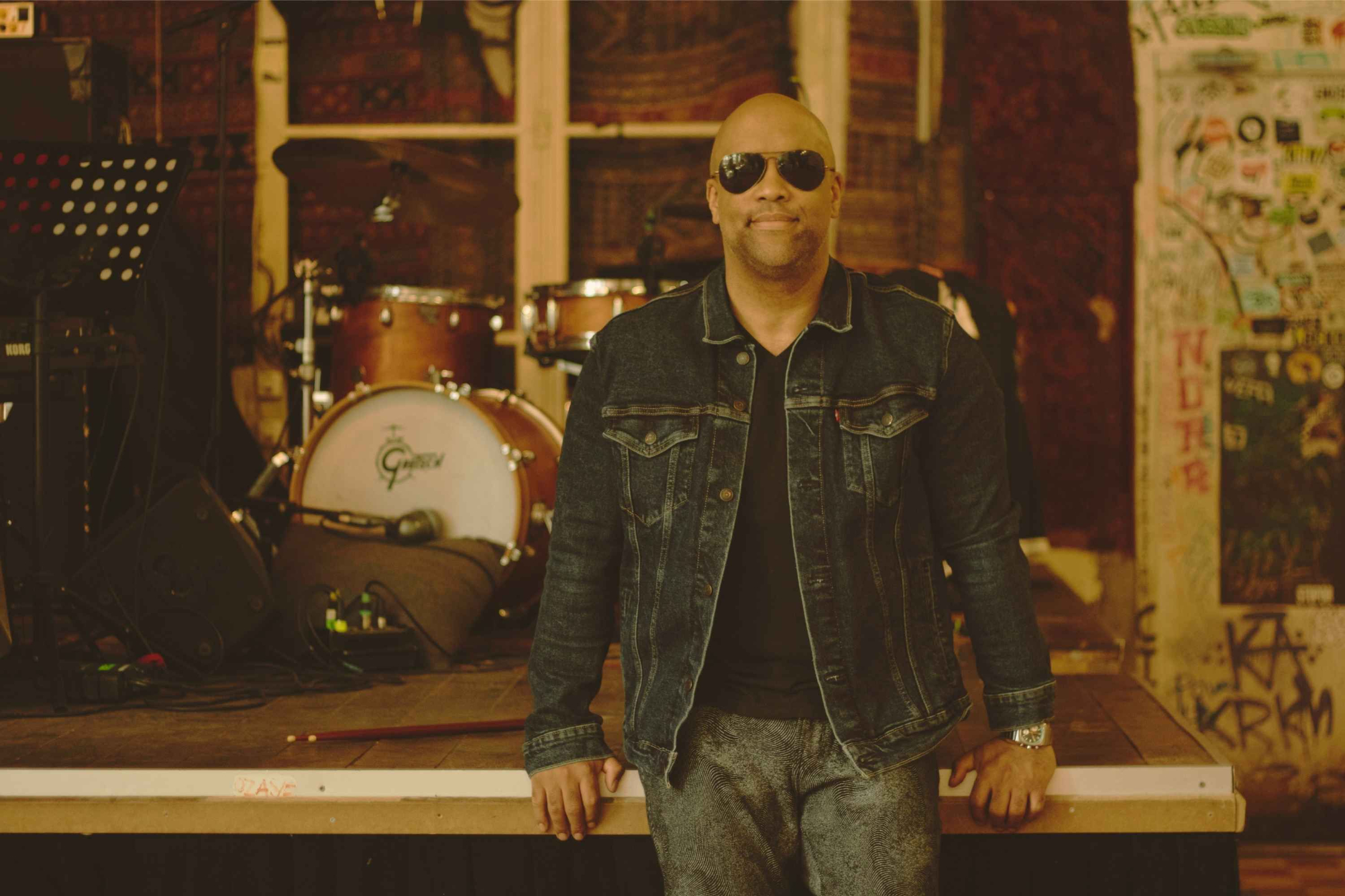 Professional drummer Mike Veny sits confidently on the edge of a stage in front of a drum kit, wearing sunglasses and a denim jacket in an intimate music venue setting.