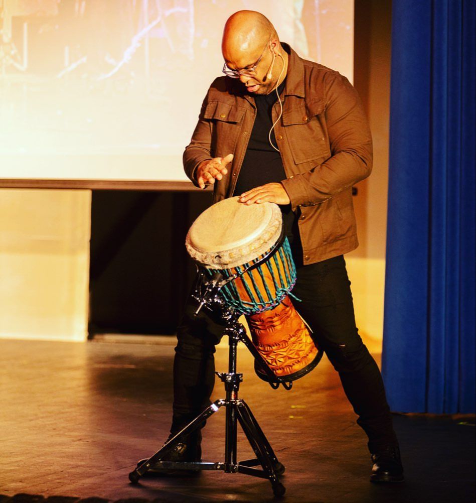 Mike Veny, a mental health speaker for college students, plays a Wula djembe during an interactive presentation. Wearing glasses, a brown jacket, and black clothing, he performs under warm stage lighting with a projection screen and blue curtain behind him.