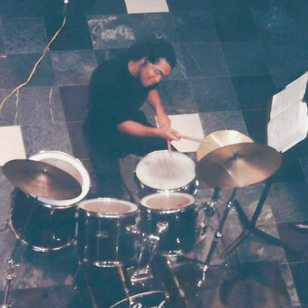 Mike Veny at 18 years old playing a Pearl drum set, photographed from above while striking the snare drum with wooden drumsticks. He is wearing a dark shirt and playing on a tiled floor, with cymbals, toms, a music stand, and sheet music visible around the drum kit.