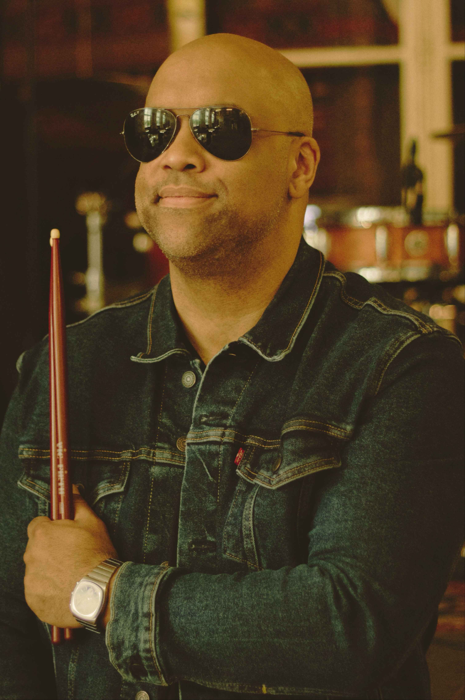 Professional drummer Mike Veny poses for a close-up portrait wearing sunglasses and a denim jacket while holding drumsticks, with a softly blurred drum kit in the background.