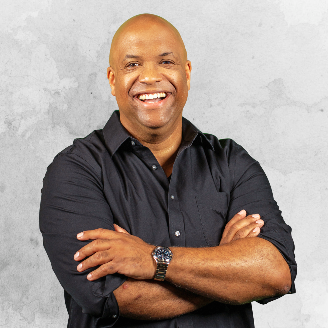 Mike Veny smiles confidently with his arms crossed while wearing a black button-down shirt and dark wristwatch. He stands against a light gray textured background in a polished professional portrait.