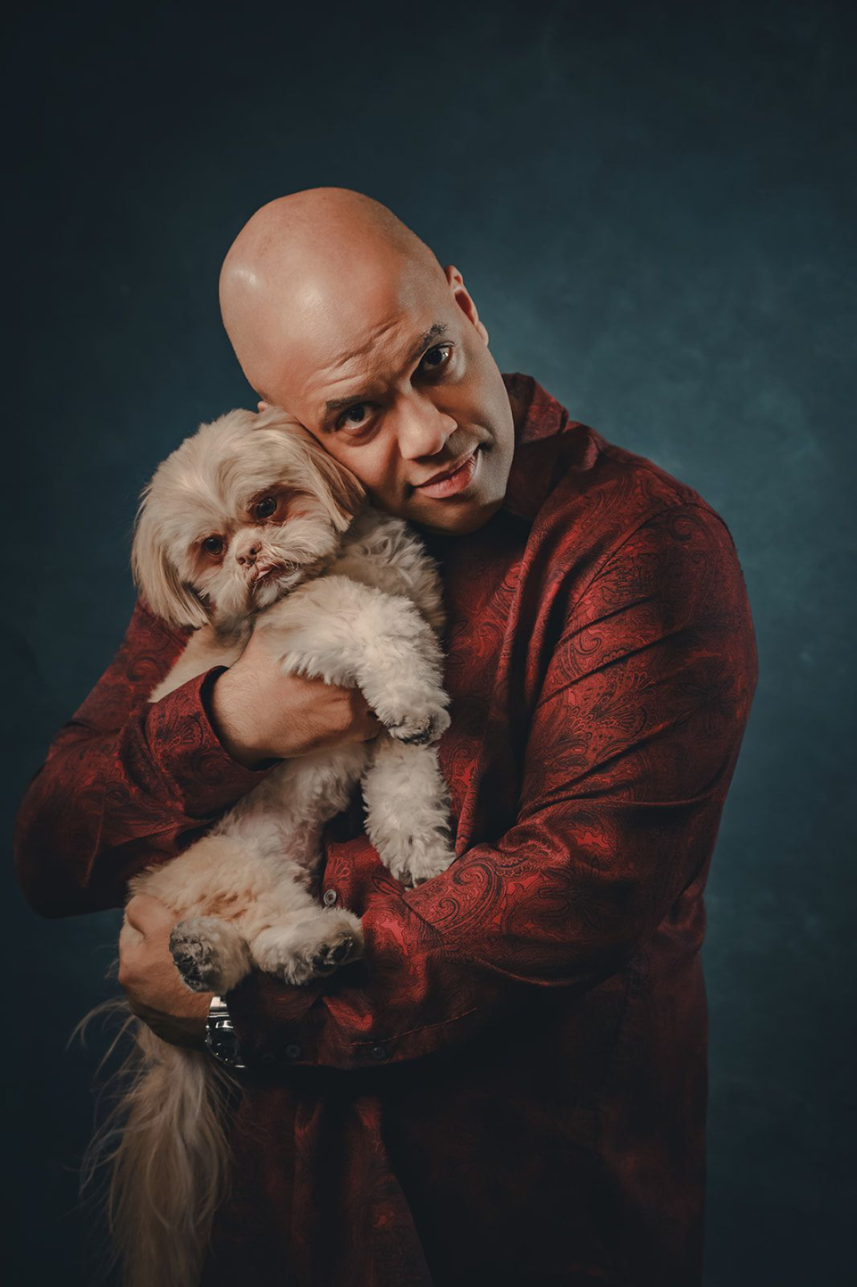 Mike Veny poses in a studio portrait while warmly holding his small white Shih Tzu, Pippin, close to his chest. Mike is wearing a rich red patterned shirt and looking directly at the camera with a gentle, affectionate expression. Pippin rests calmly in his arms, facing forward, against a dark teal textured backdrop with dramatic studio lighting that creates an intimate, polished, and emotionally warm portrait.