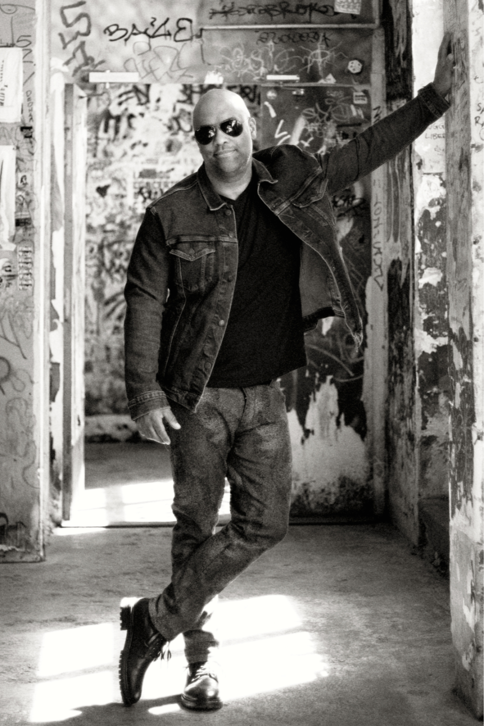 Black-and-white portrait of Mike Veny, a patient advocate and wellness influencer, standing in a graffiti-covered urban hallway wearing sunglasses, a denim jacket, jeans, and boots, with one arm resting against the wall in a confident, artistic pose.