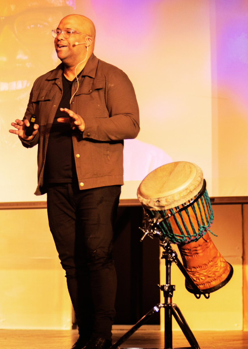 Mike Veny, an inspirational mental health keynote speaker, stands onstage beside a colorful African djembe drum mounted on a stand. Wearing glasses, a black shirt, black jacket, and black pants, he smiles while speaking into a headset microphone and holding a presentation remote. His hands are raised expressively as he addresses the audience. Warm orange, yellow, pink, and purple stage lighting fills the background, creating an energetic and uplifting atmosphere.