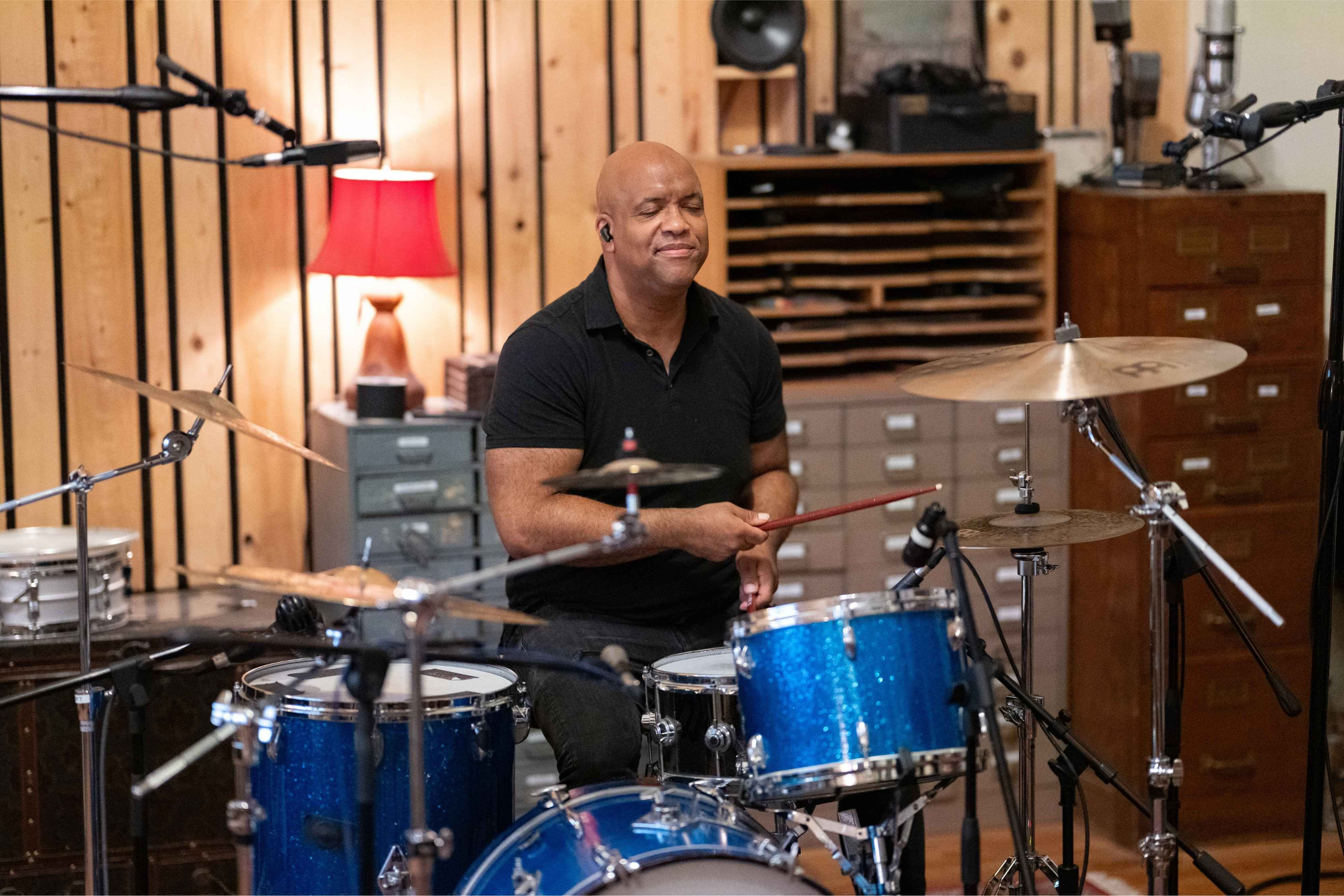 Mike Veny, one of the drummers in NYC, smiles while playing a blue drum kit in a warm recording studio surrounded by cymbals, microphones, and music equipment.