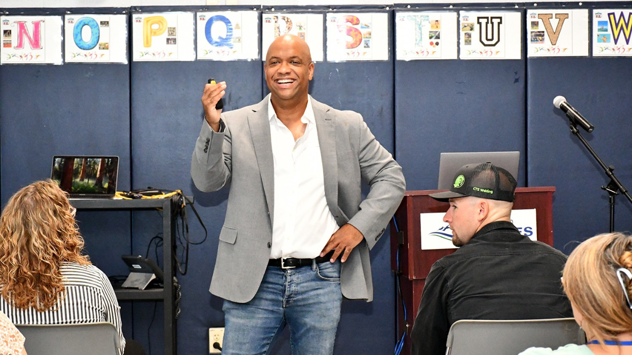 Workplace mental health speaker Mike Veny smiles while leading an interactive presentation for a seated audience in a classroom-style setting, holding a presentation remote beside a microphone and laptop.