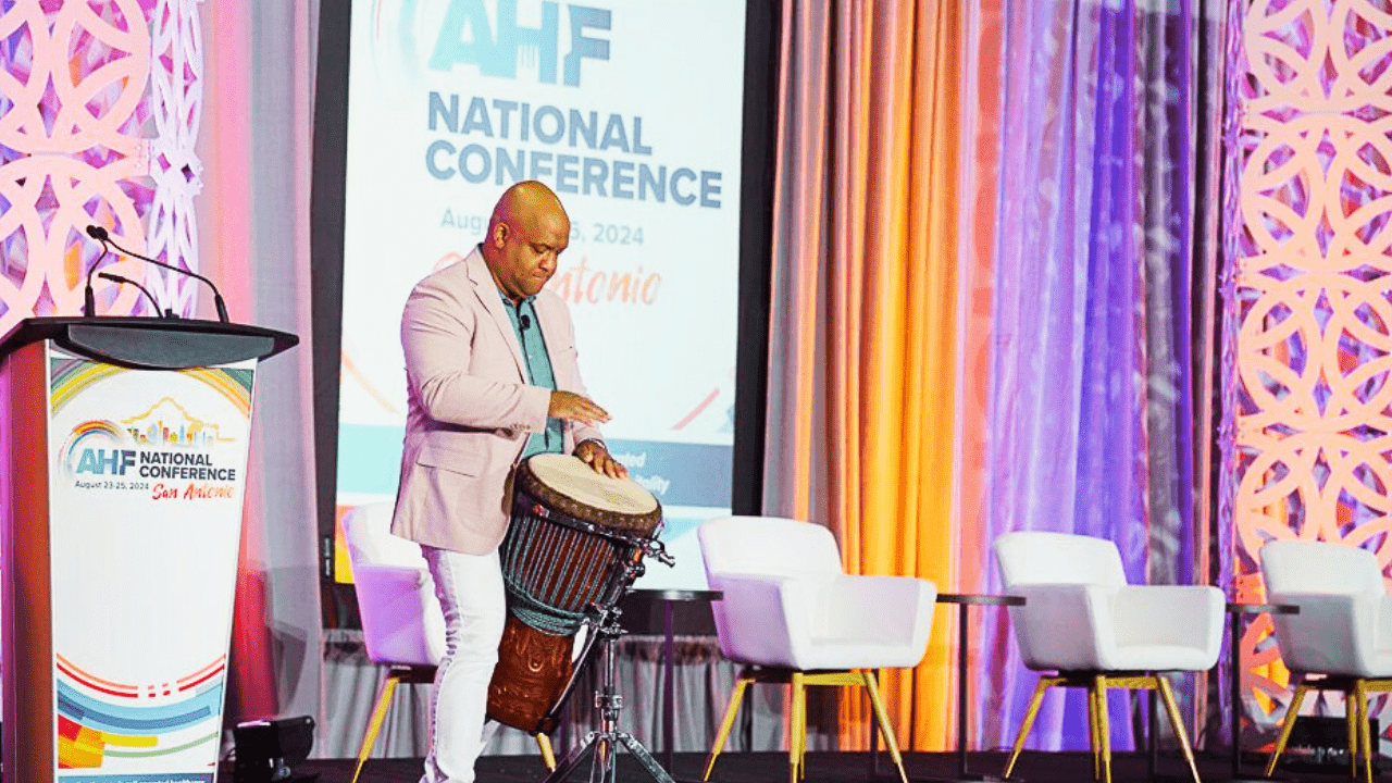 Healthcare keynote speaker Mike Veny plays a djembe onstage during the AHF National Conference in San Antonio, surrounded by colorful event lighting.