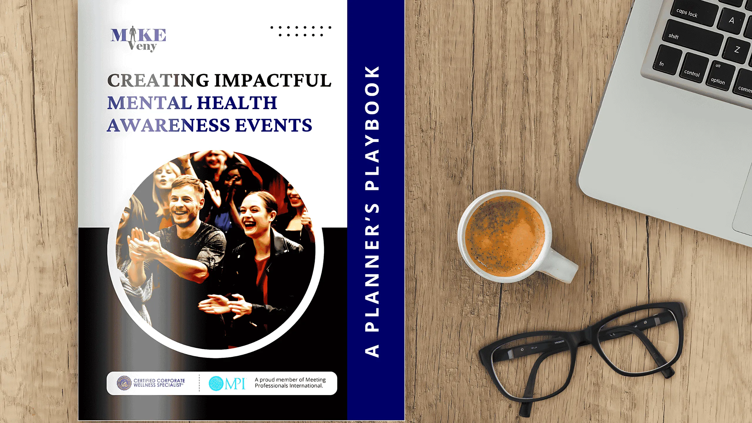 Promotional graphic for Mike Veny’s “Creating Impactful Mental Health Awareness Events: A Planner’s Playbook.” The guide is displayed on a wooden desk beside a cup of coffee, black eyeglasses, and the corner of a laptop. The cover features a smiling audience applauding at an event, the Mike Veny logo, and logos for Certified Corporate Wellness Specialist and Meeting Professionals International.