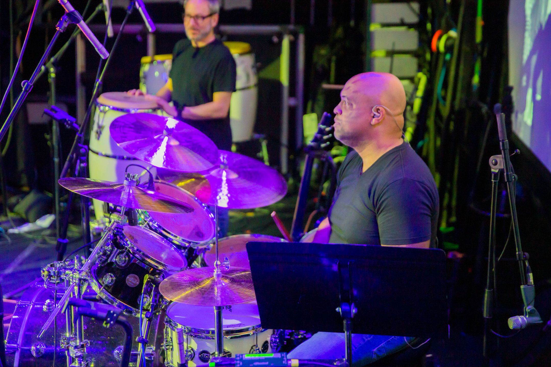 Live drummer Mike Veny plays a drum kit during a concert performance, surrounded by cymbals, microphones, stage lighting, and another percussionist in the background.