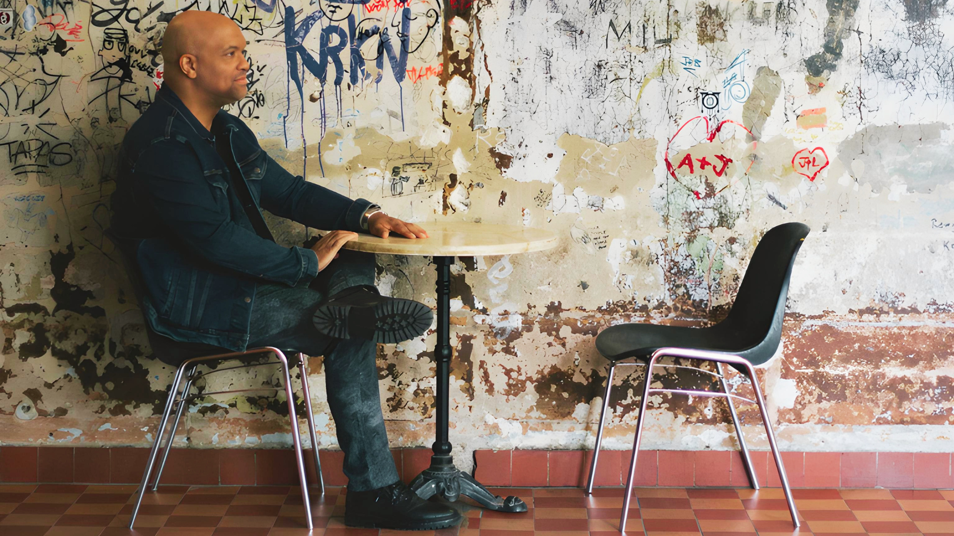 Mike Veny sits thoughtfully at a small round café table with an empty chair across from him, suggesting an intimate conversation for his Coffee With Mike podcast. He wears a dark denim jacket and jeans against a textured, graffiti-covered wall in a creative urban setting.