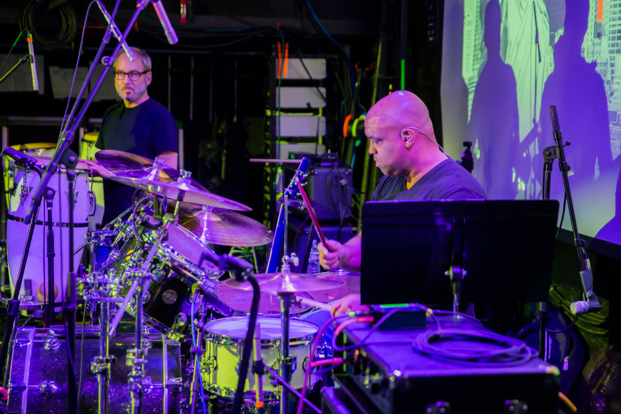 Live band drummer Mike Veny performs on stage in New York City with a full drum kit, microphones, percussion, and colorful purple and green concert lighting.