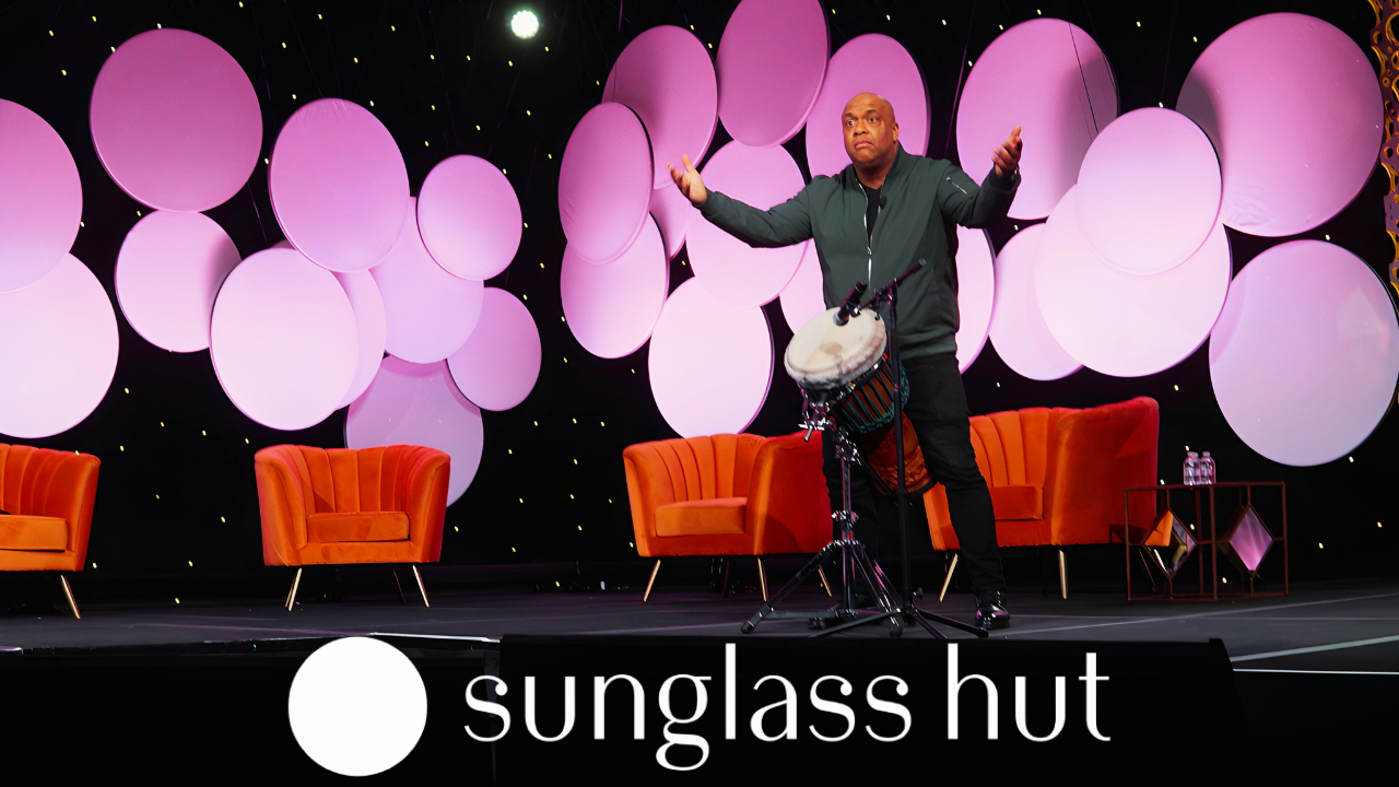 Mike Veny, mental health keynote and motivational speaker and professional drummer, stands behind a djembe with his arms extended while engaging an audience on a colorful Sunglass Hut event stage featuring orange seating and illuminated pink circular panels.