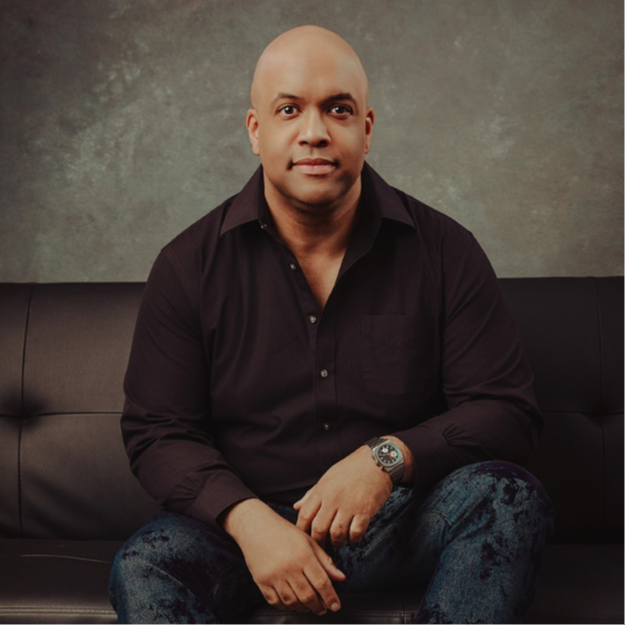 Portrait of Mike Veny seated on a dark, tufted leather couch against a textured gray studio backdrop. He is wearing a black short-sleeve button-up shirt, dark jeans, and a wristwatch, with his hands resting naturally in front of him. The image has a professional, calm, and approachable tone, presenting Mike Veny as a confident mental health speaker, professional drummer, and Meeting Professionals International member.