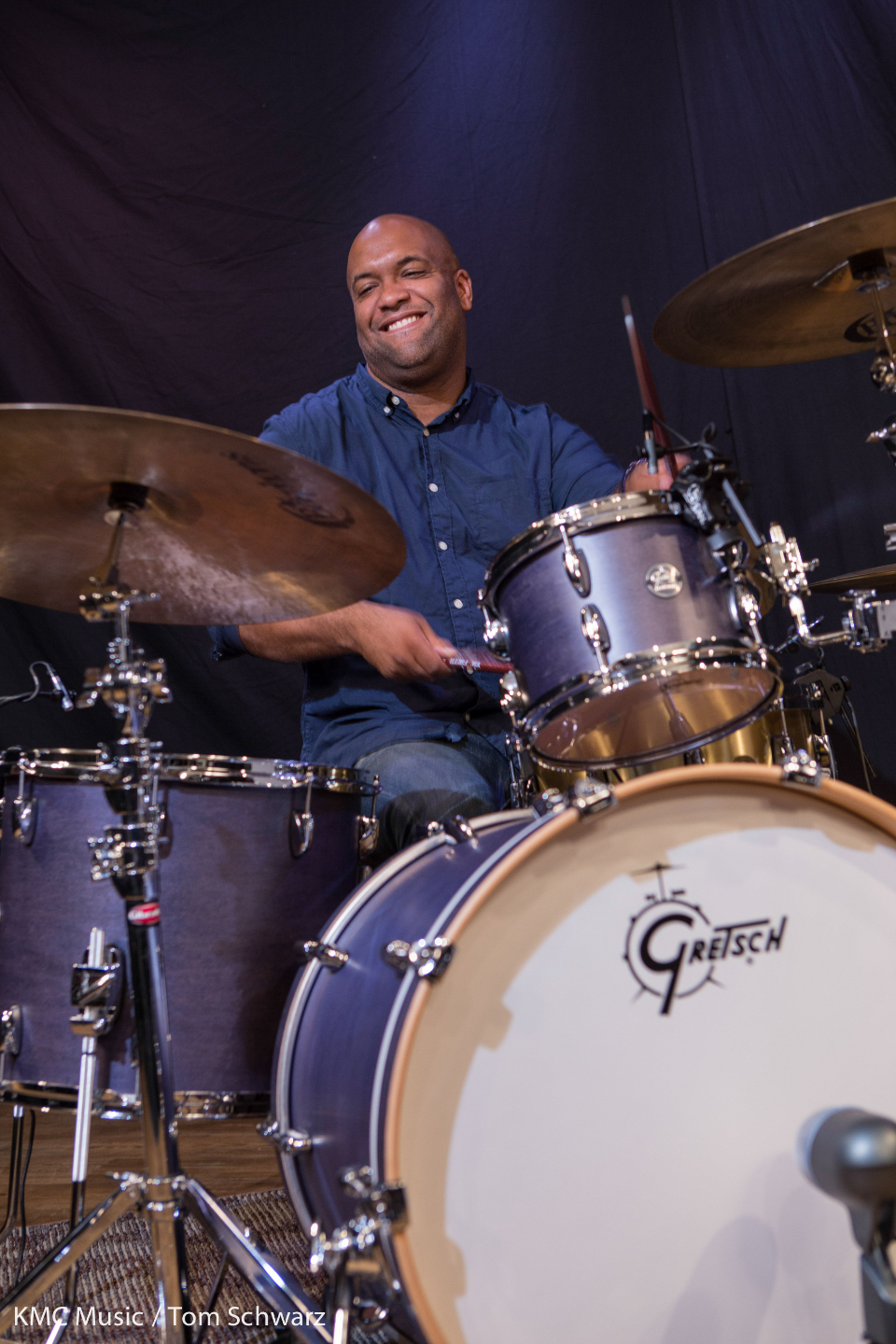 Mike Veny smiles while performing on a Gretsch drum kit, captured mid-action behind the cymbals and drums. He is wearing a dark blue button-down shirt and holding drumsticks as he plays in a studio setting with a dark backdrop, conveying the energy and joy of a professional live drummer.