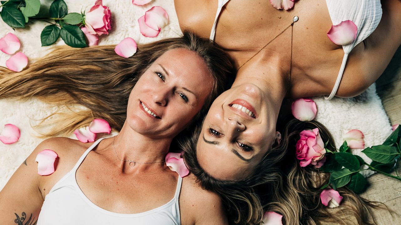 Two women lying side by side surrounded by pink rose petals, smiling softly in a calm and intimate wellness-inspired setting.