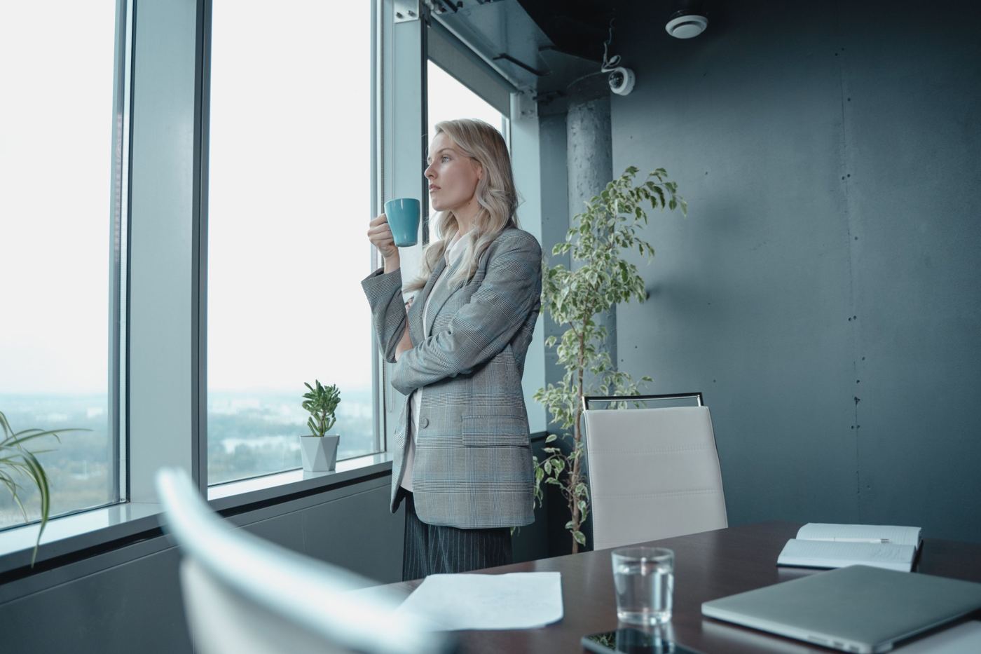 A woman in a blazer looking out an office window with a coffee mug
