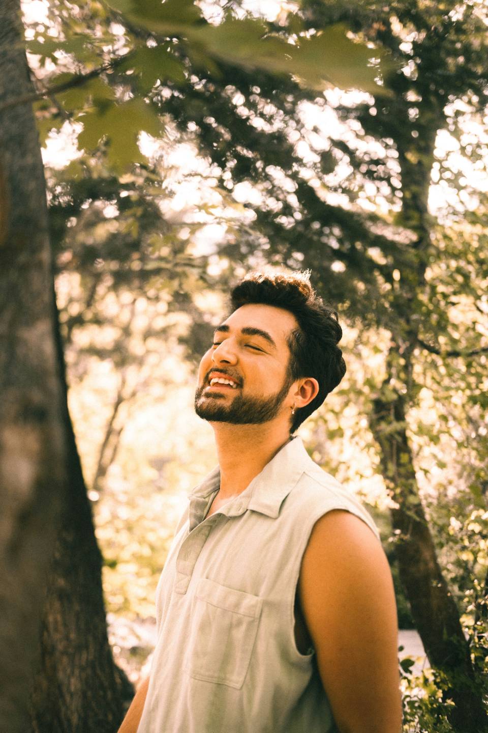 A man standing among sunlit trees, smiling with his eyes closed