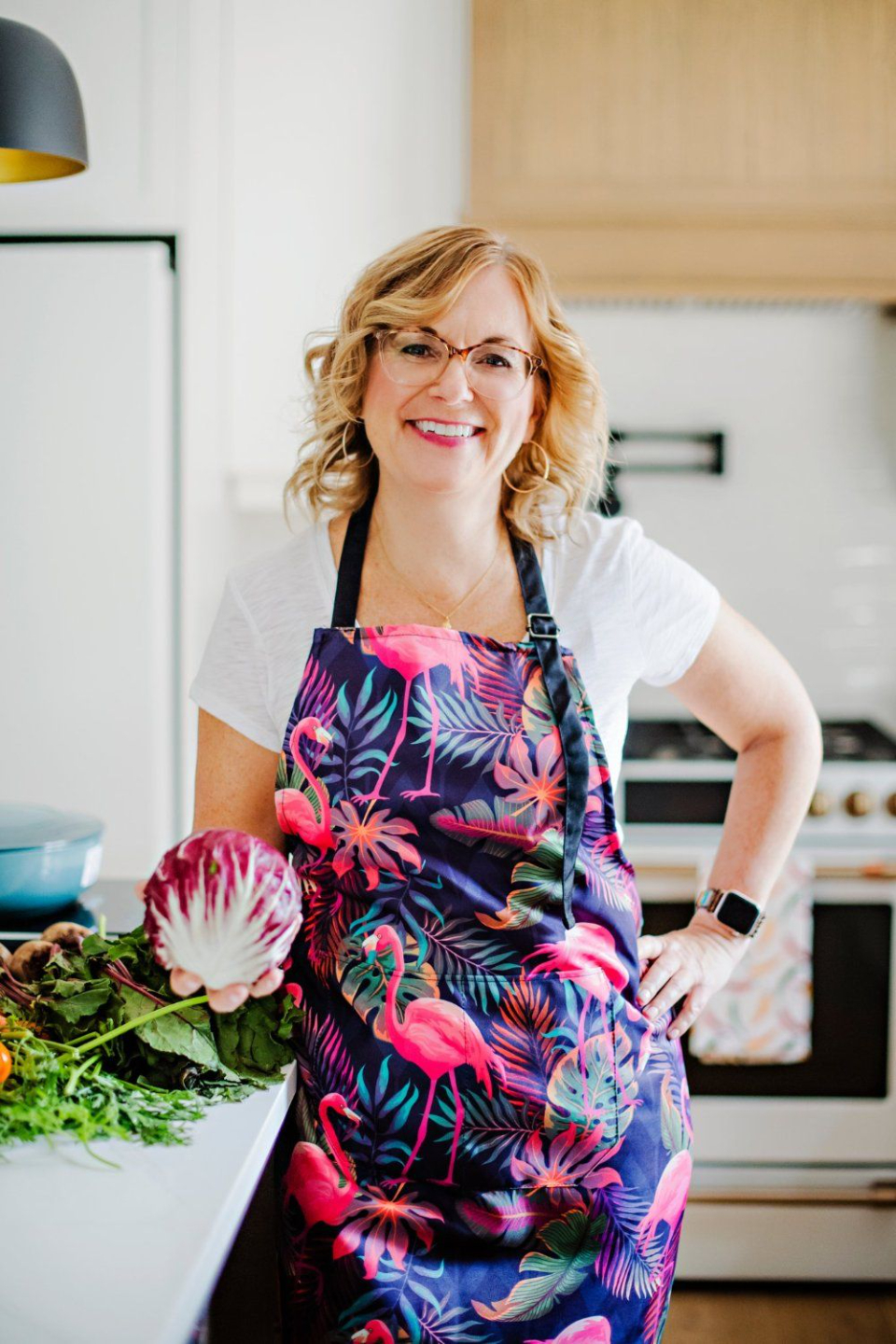 Lynne Zimmerman in a colorful flamingo-print apron, holding fresh cabbage and greens in her kitchen