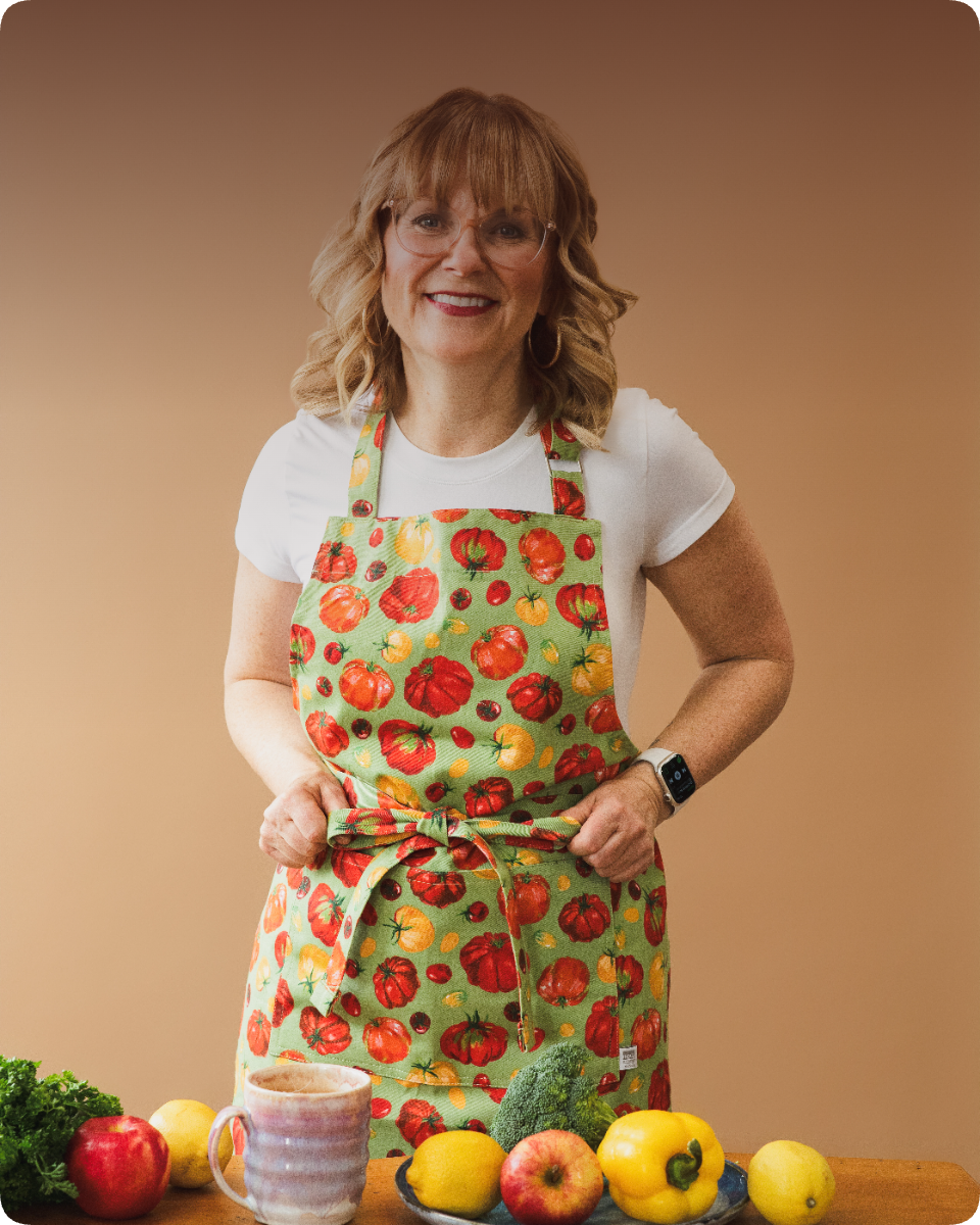 Lynne Zimmerman standing at a kitchen counter in a tomato-print apron, surrounded by fresh produce