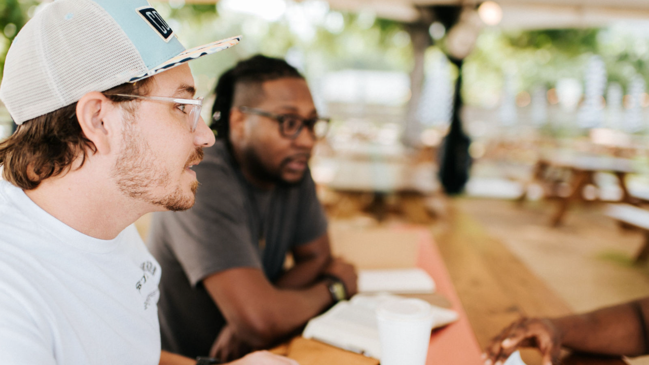 A diverse group of men and women sit together in a warm living room — some on the floor, some on the sofa — sharing quiet conversation as they explore how to heal emotional wounds.