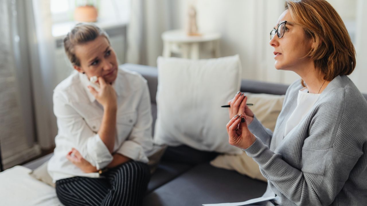 Two women sit on opposite ends of a cream linen sofa in a warm living room, turned toward each other in quiet conversation — one holding a golden mustard mug with both hands as she listens, the other with her mug on the walnut side table beside her as she speaks softly.