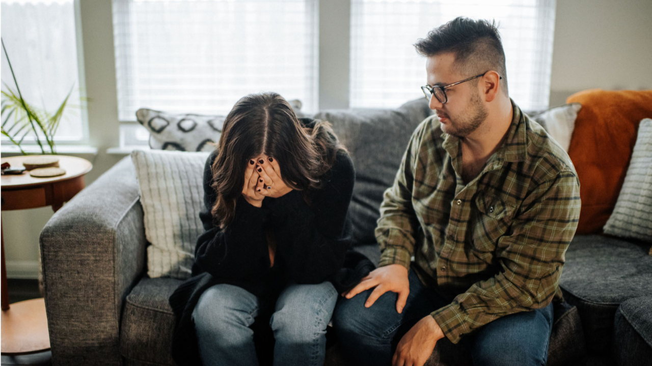 A young man sits slouched in a cream boucle chair, hand pressed to his temple in quiet frustration, a book open on his lap and a coffee mug beside him — the look of someone exhausted from trying to think his way out of pain.