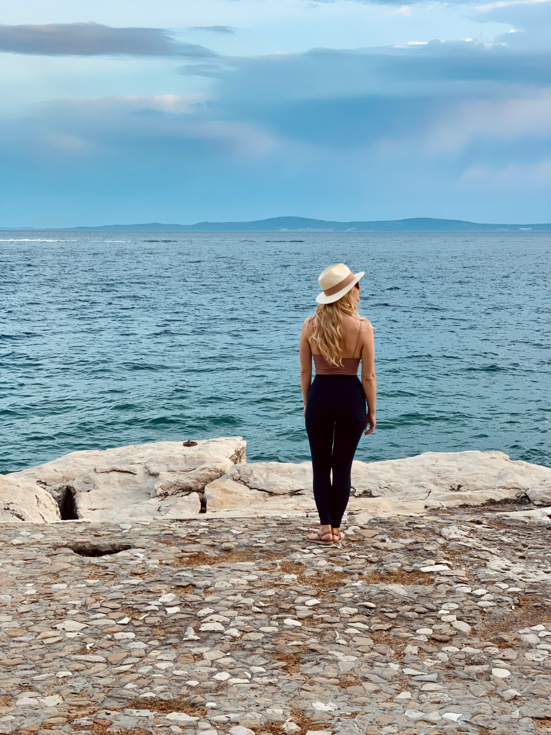A photo of Rebecca Michelle a personal mindfulness coach from nyc who is standing by the sea and has a beige hat on.