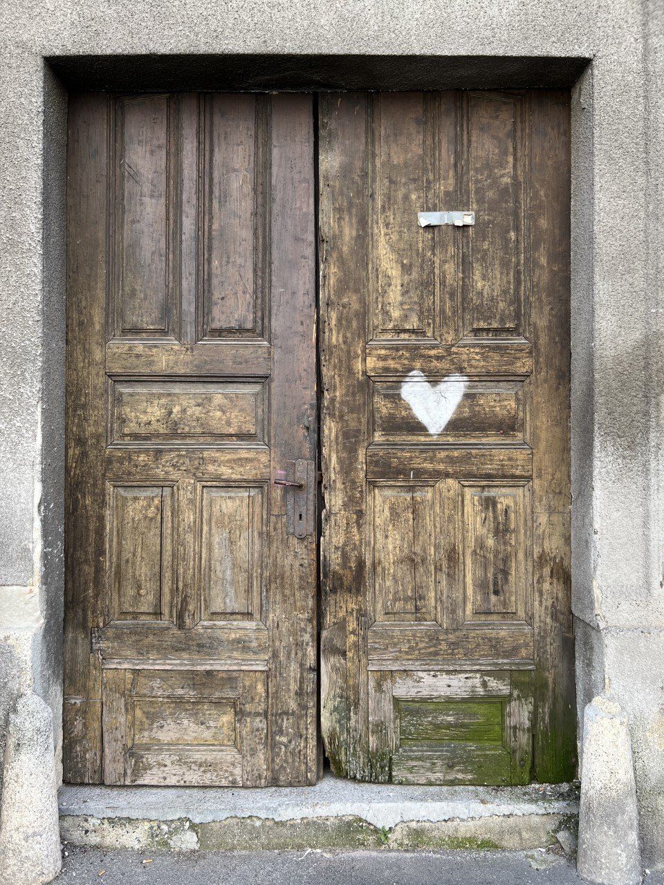 Old wooden double door with a white heart spray-painted on the right door, surrounded by a stone frame, with weathered and stained wood.