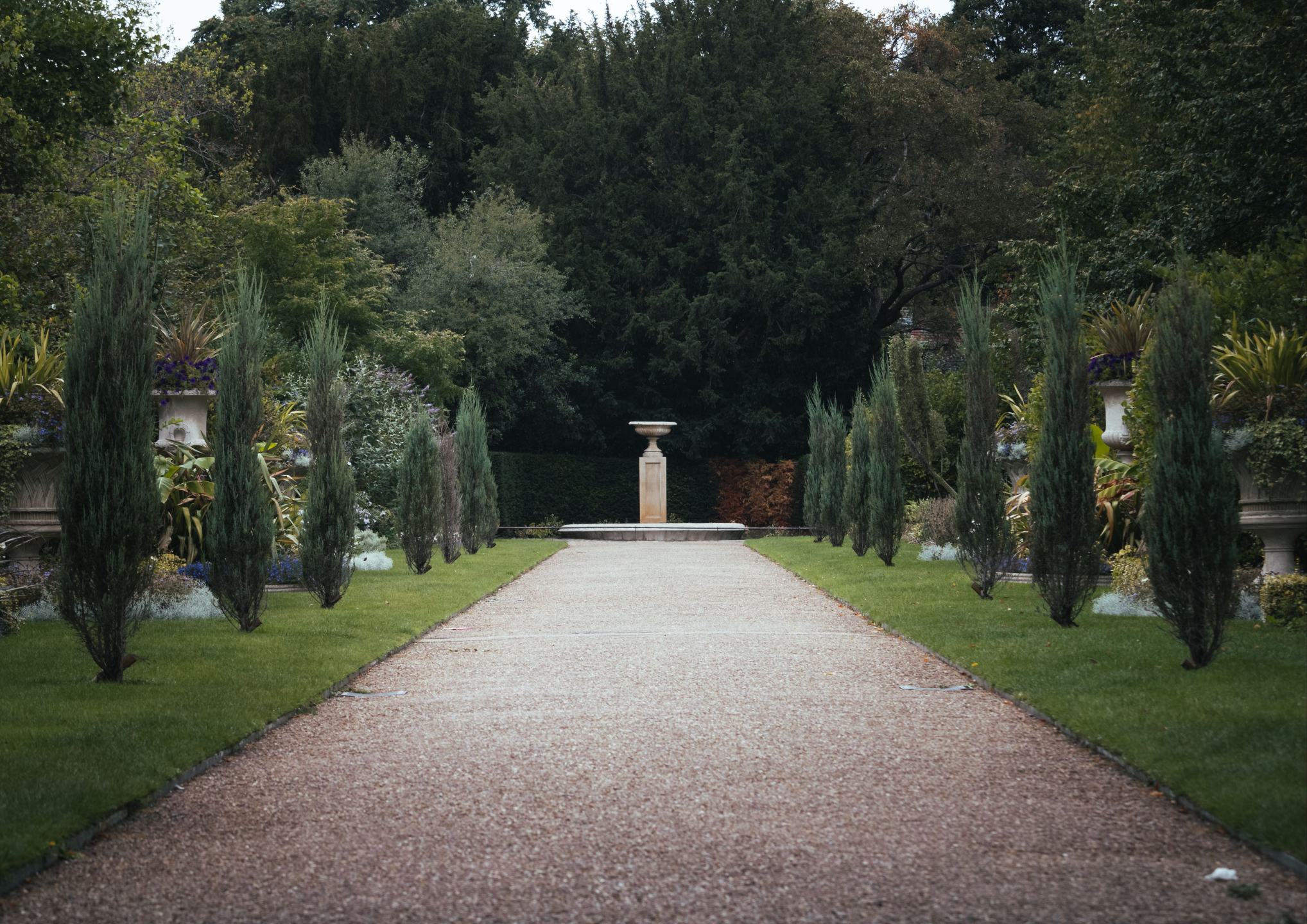A gravel walk between clipped cypresses, running toward a stone urn on its plinth