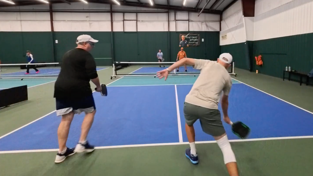 A pickleball player in a light shirt swings at the ball on an indoor court while another player in a dark shirt looks on.