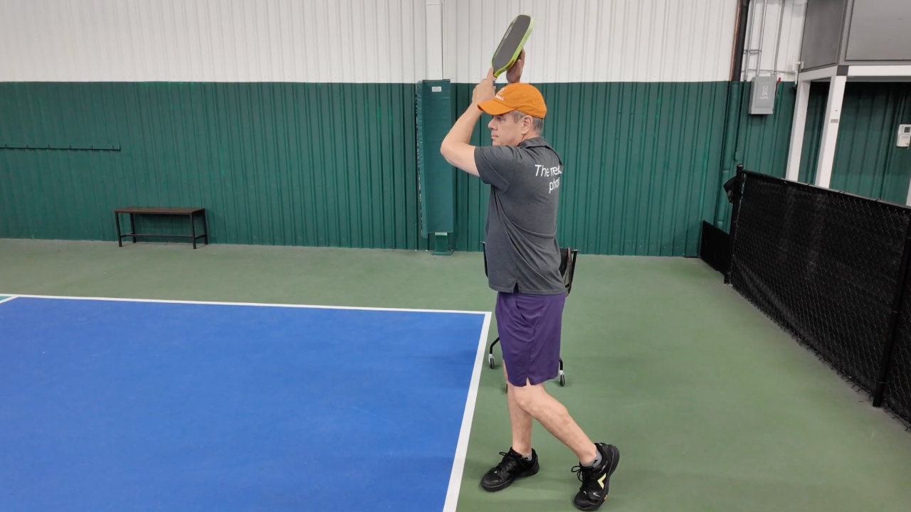 Coach Tony Roig stands beside an indoor pickleball court with his paddle raised high behind his head as he completes his serve