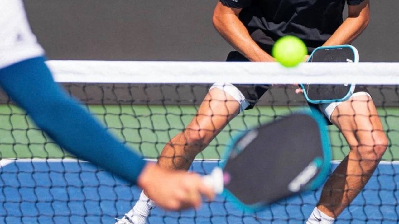 Close-up of a pickleball player at the kitchen line reaching a paddle toward a yellow pickleball, wearing a blue arm sleeve, on a blue court.