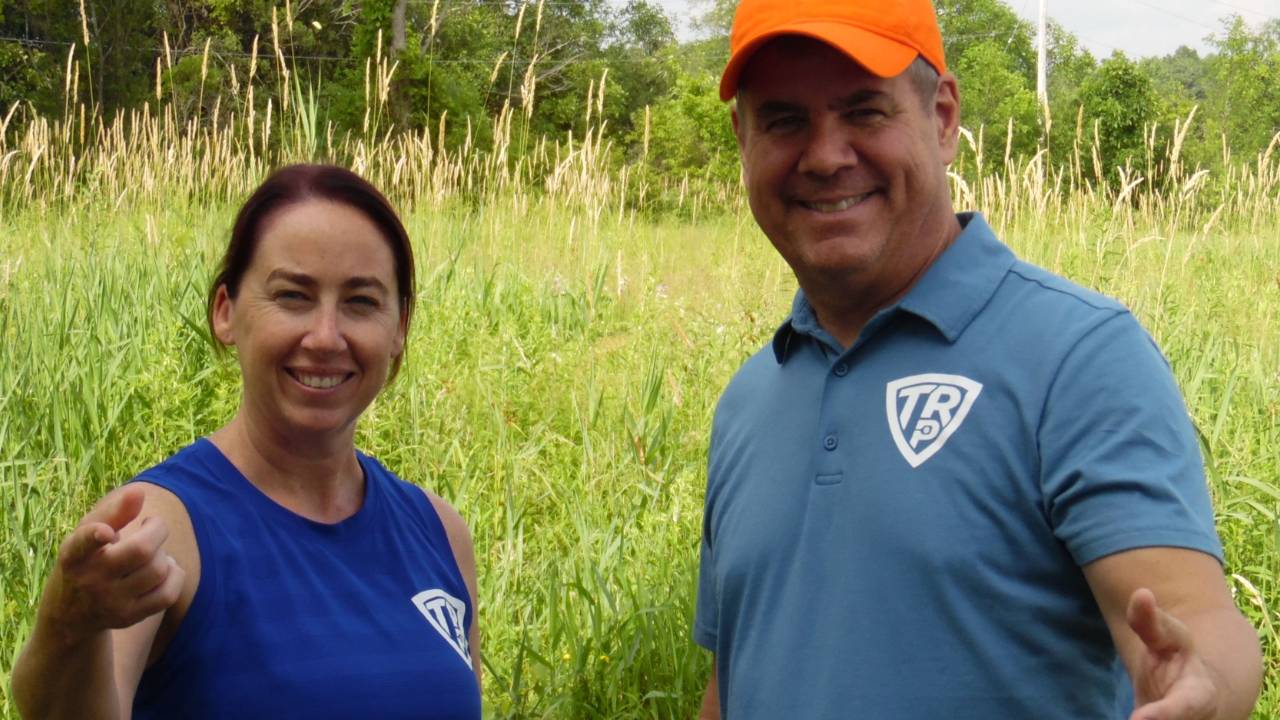 Kyleen Dye and Coach Tony Roig smiling and pointing at the camera while standing in a field of tall grass