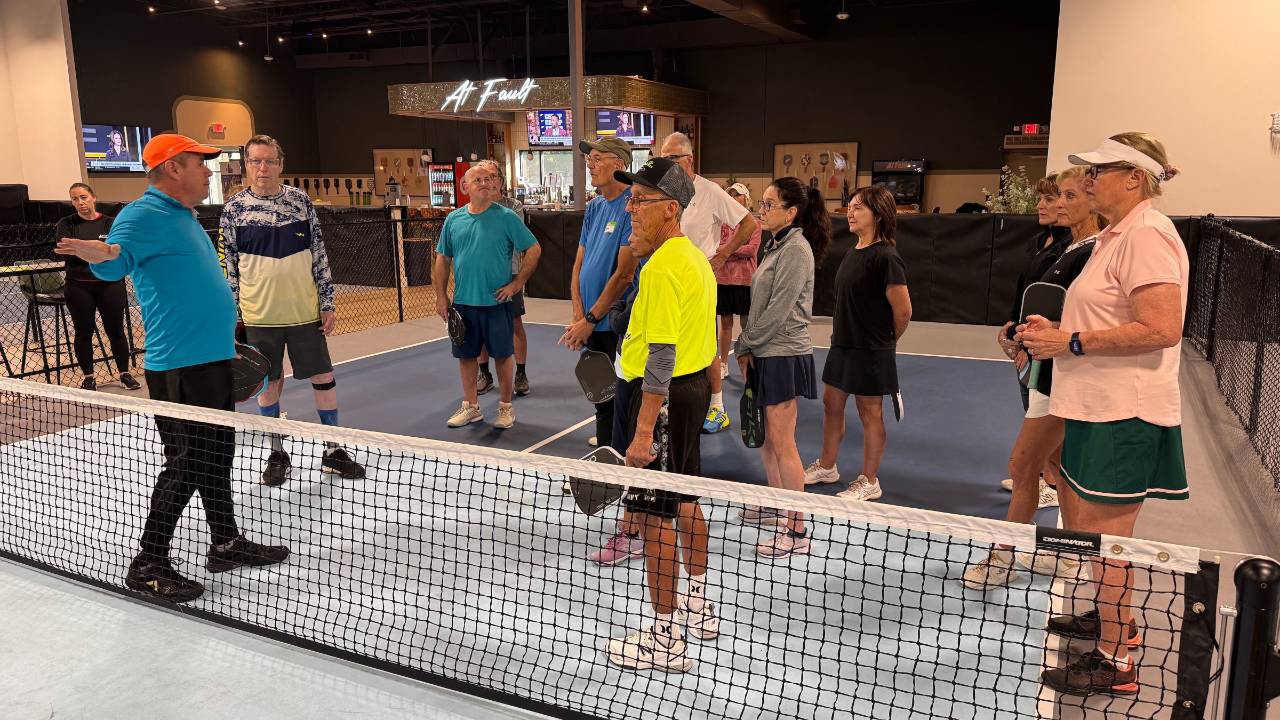 Coach Tony Roig talking with a group of adult players at the NVZ line during a pickleball camp