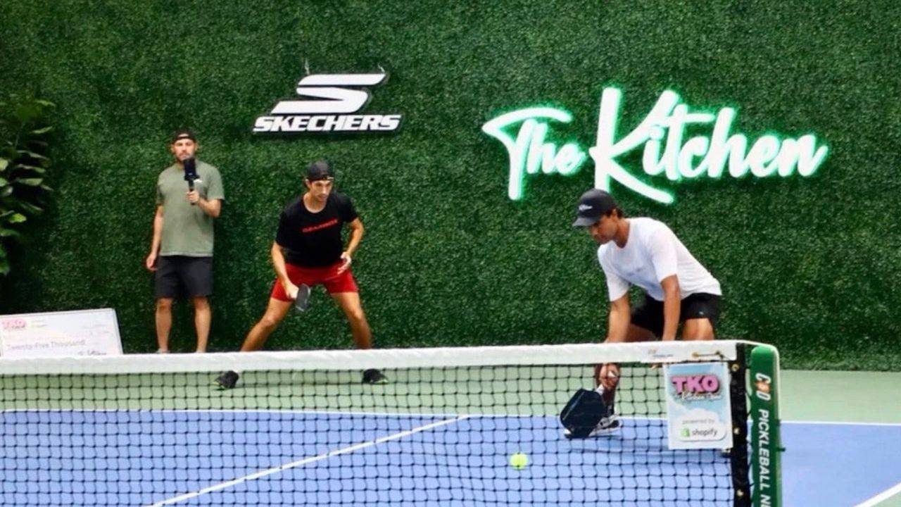 Pickleball players navigating the Transition Zone during a rally, with Skechers and The Kitchen signage on a green hedge wall behind the court.