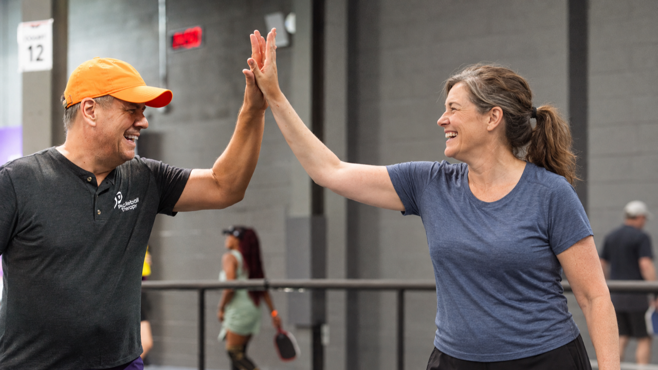 Coach Tony Roig in an orange cap high-fives a smiling player at an indoor pickleball facility