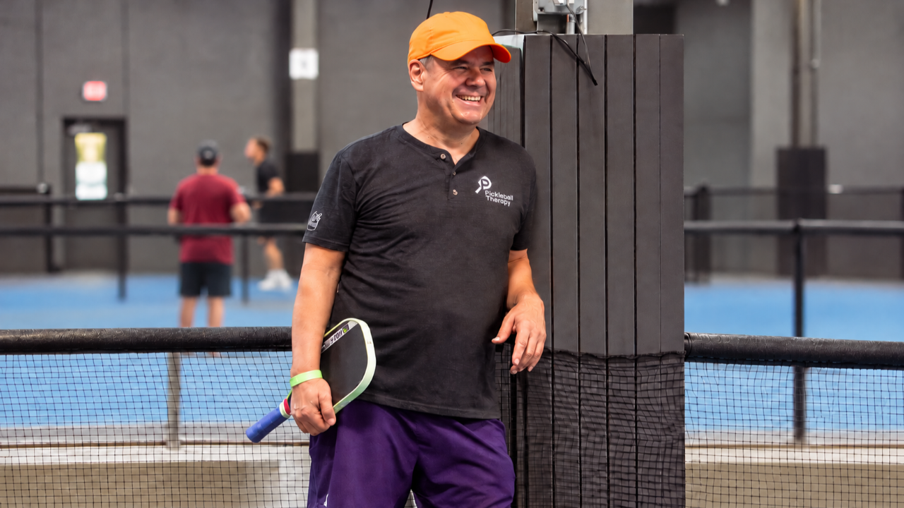 Coach Tony Roig smiling and holding a pickleball paddle at an indoor pickleball facility